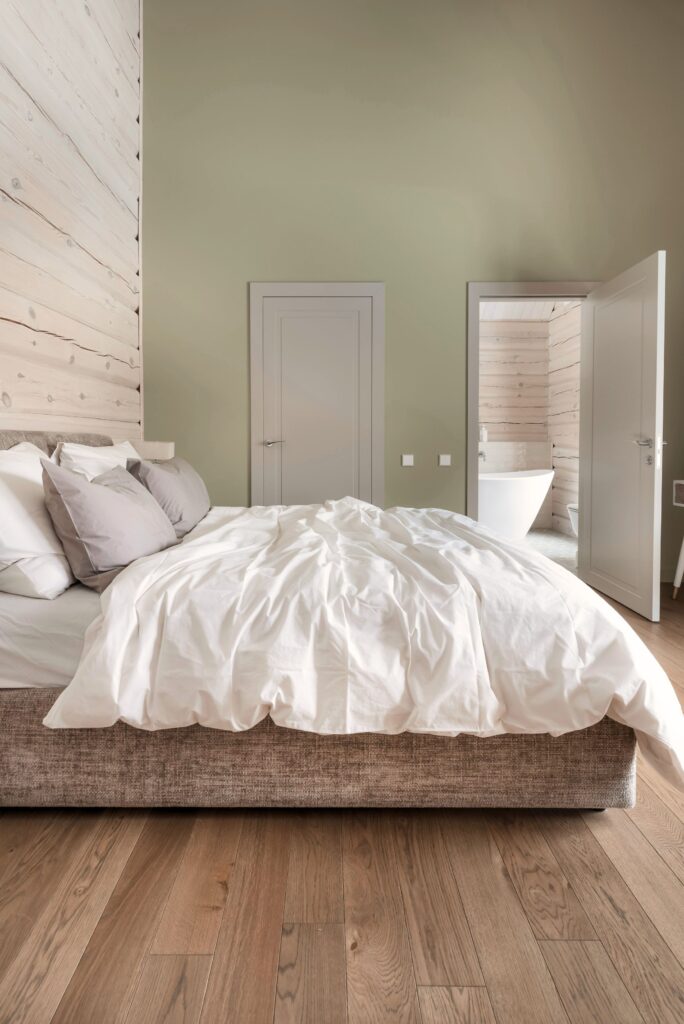 Tamsiai rudos grindys miegamajame, dvigulė lova, balta patalynė ir šviesios sienos